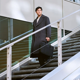 Man walking down outdoor stairs carrying a black laptop bag, showing the bag styled as a modern business briefcase for commuting, office wear, or professional travel.