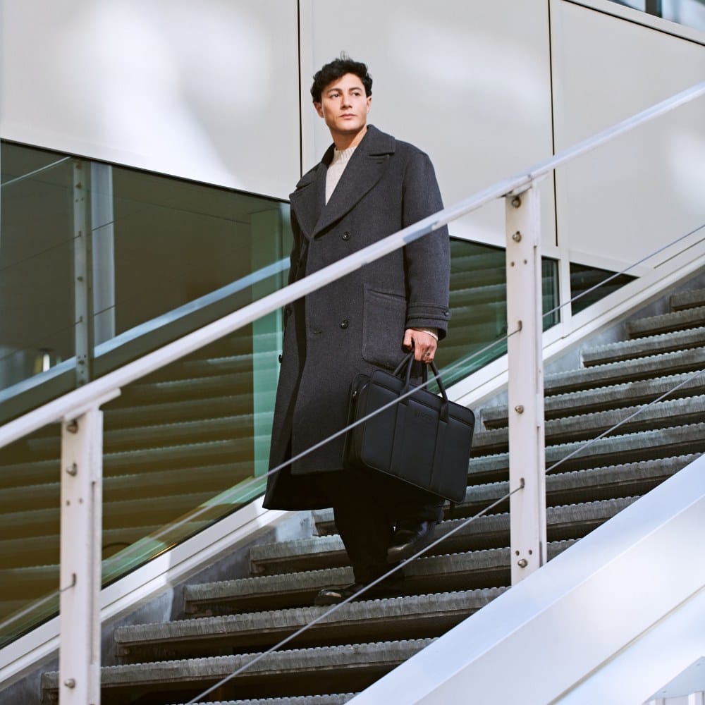 Man walking down outdoor stairs carrying a black laptop bag, showing the bag styled as a modern business briefcase for commuting, office wear, or professional travel.