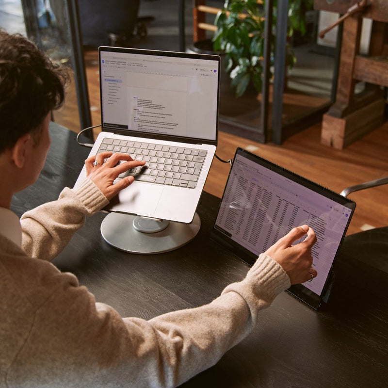 Person using a silver laptop stand at a desk with the laptop raised to eye level beside a portable monitor, creating an ergonomic dual-screen workspace for productivity.