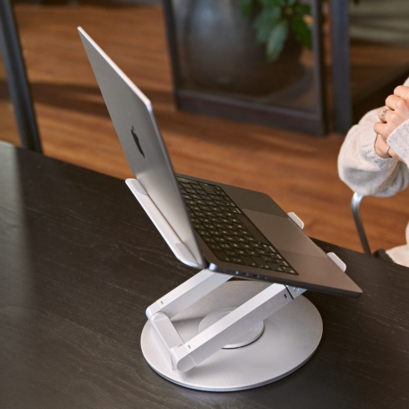 Side view of a silver laptop stand with an open laptop, showing the adjustable arms, elevated typing angle, and round rotating base on a dark wood desk.