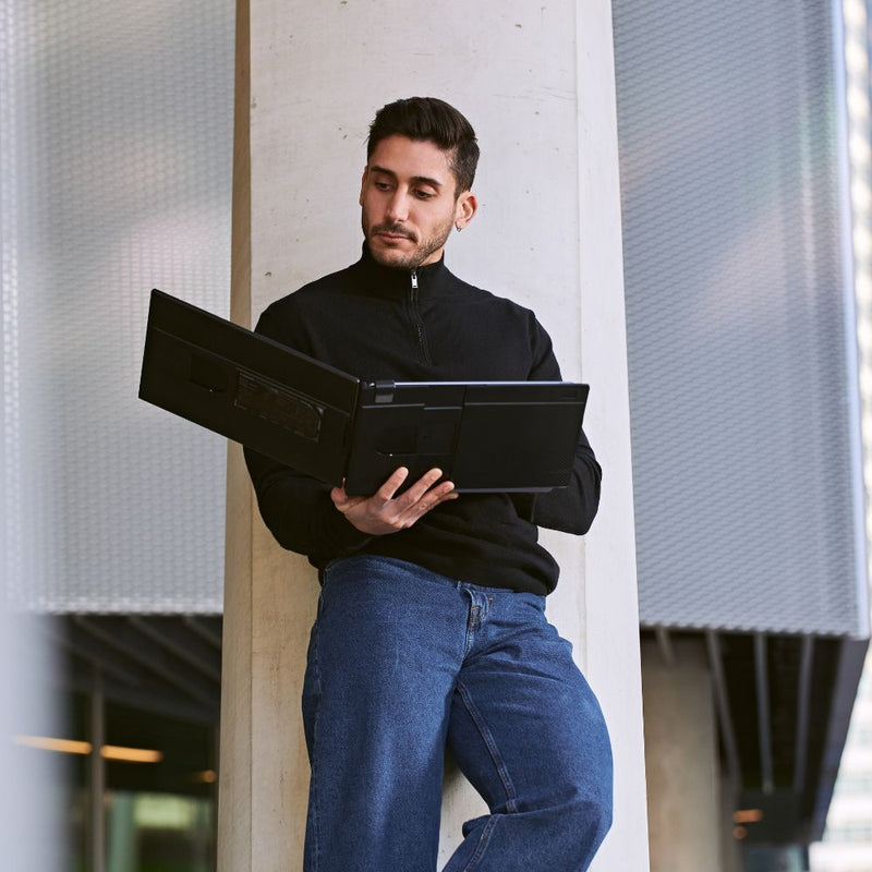 Person leaning against a concrete pillar while holding an open portable monitor with a dual-screen setup, showing the slim black design and fold-out screens for mobile productivity.