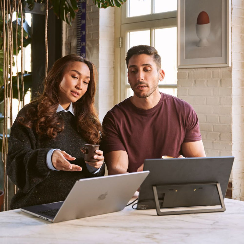 Two coworkers sitting at a table with a laptop and a portable monitor, discussing work in a bright indoor space, showing a collaborative dual-screen setup for meetings and shared productivity.