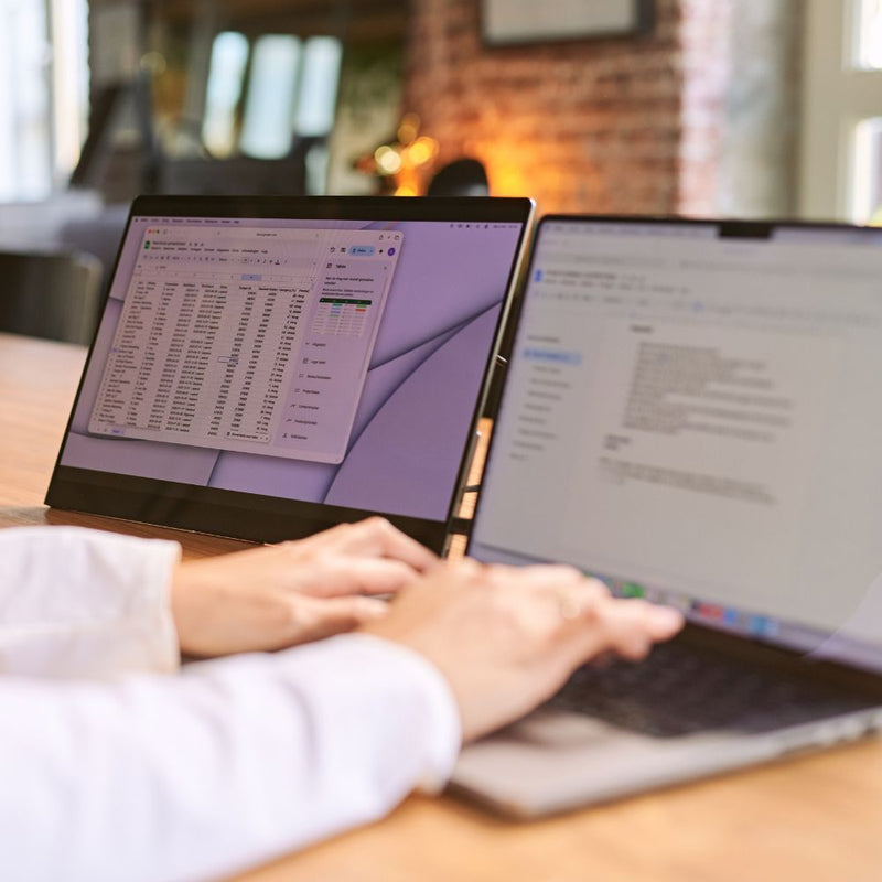 Close-up of hands typing on a laptop beside a portable monitor, with a spreadsheet open on the external display and a document on the laptop for an efficient dual-screen workspace.