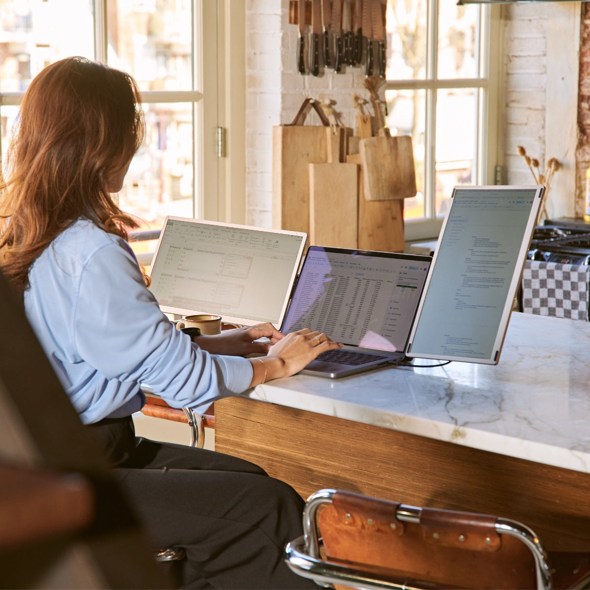 Portable monitor triple-screen workstation in a bright kitchen, with a woman using spreadsheets and documents across three displays.