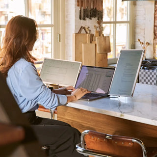 Woman working with a triple-screen portable monitor setup in a bright kitchen, viewing spreadsheets and documents across three displays.