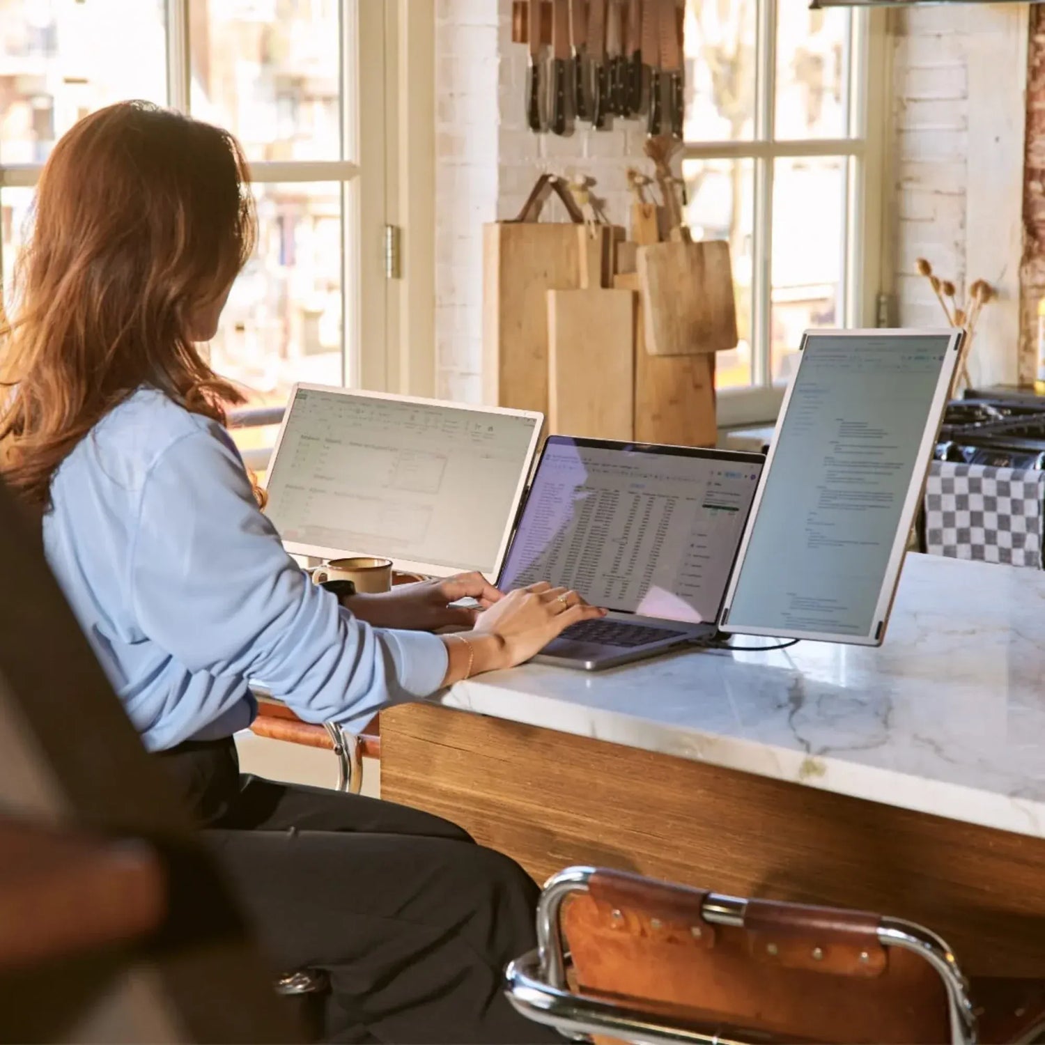 Woman working with a triple-screen portable monitor setup in a bright kitchen, viewing spreadsheets and documents across three displays.