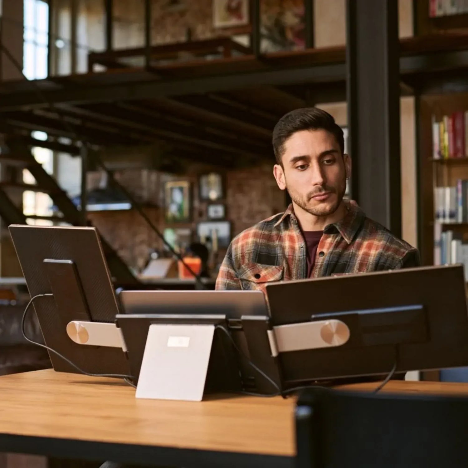 Man working behind a laptop with two portable monitor side screens supported by adjustable rear stands on a wooden desk.