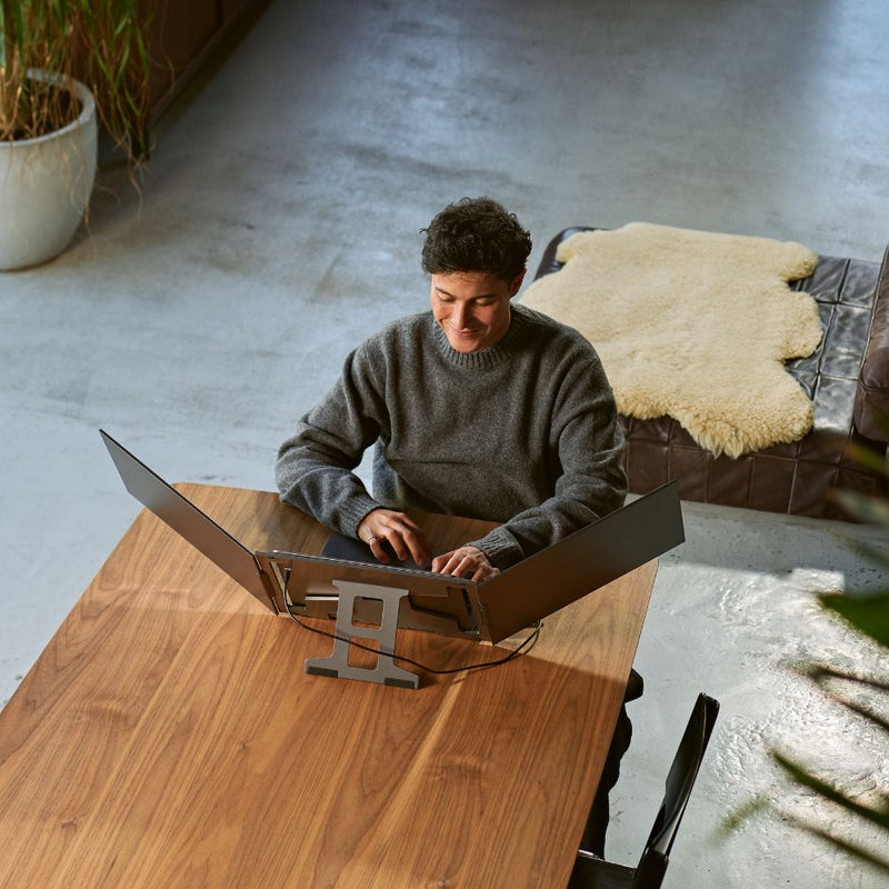 Overhead view of a person working at a wooden table with a portable monitor triple-screen setup, showing two side displays extended from a central laptop for multitasking and productivity.
