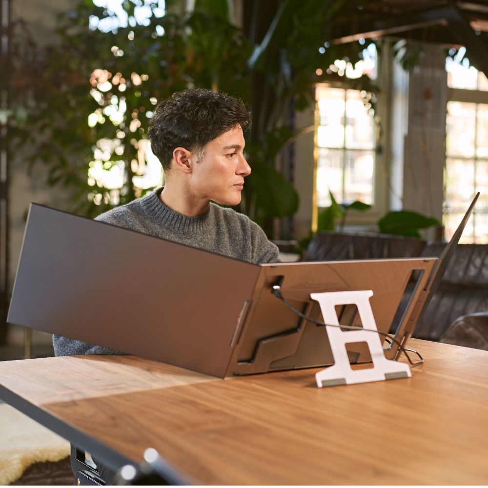 Side view of a person using a portable monitor with a triple-screen workstation, featuring two wide side panels connected to a laptop on a stand in a bright indoor workspace.