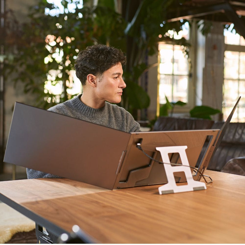 Side view of a person using a portable monitor with a triple-screen workstation, featuring two wide side panels connected to a laptop on a stand in a bright indoor workspace.